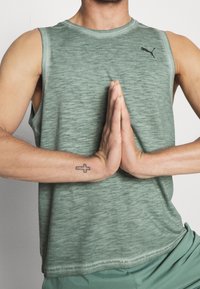 Jasnozielony, bezrękawnik sportowy z subtelną fakturą i logo Pumy na piersi. Ręce są ułożone w geście modlitwy.