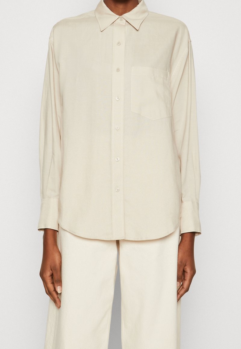 Personne portant une chemise ample crème à boutons avec poche poitrine et un pantalon large assorti sur un fond uni.