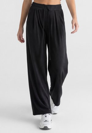 Person trägt lockere schwarze, weit geschnittene Hose mit Kordelzug in der Taille und weißen Sportsneakers und geht nach vorne.