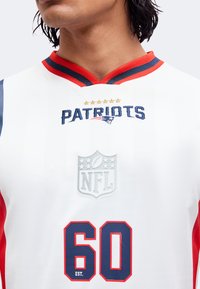 Άνδρας που φοράει λευκή φανέλα των Patriots NFL με κόκκινο και μπλε γιακά και τον αριθμό 60 στο μπροστινό μέρος.