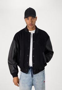 Veste varsity noire avec corps en laine et manches en cuir. Présente un devant à boutons, un col côtelé et un logo sur la poitrine. Portée avec un jean en denim clair.