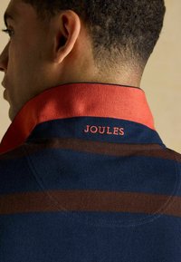 Jūras zils polo krekls ar sarkanu apkakli, rotāts ar horizontālām brūnām svītrām. "JOULES" ir izšūti korallos uz apkakles. Tekstūras audums.