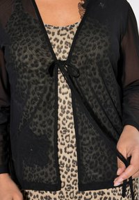 Cardigan trasparente nero con chiusura a fiocco, indossato sopra un top beige con stampa leopardata. Presenta una texture morbida e dettagli a contrasto nei motivi.