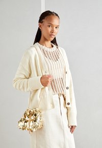 Kremowy dziergany kardigan o prążkowanej fakturze, zestawiony z beżowym topem szydełkowym. Trzyma małą złotą torebkę typu bucket ozdobioną cekinami. Gładka, lekka tkanina.