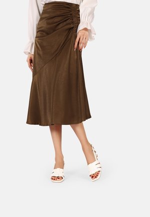Vrouw draagt een bruine geplooide midi-rok met een witte blouse met lange mouwen en witte open teen sandalen met hak.