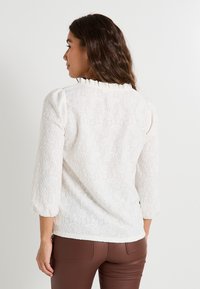 Blusa blanca con un patrón de paisley texturizado, mangas de tres cuartos abullonadas y un escote con volantes, combinada con pantalones ajustados marrones.