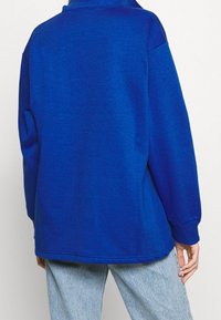 Överdimensionerad blå sweatshirt med hög krage, långa ärmar, ribbade muddar och detaljer i fållen. Mjuk textur, lämplig för avslappnad användning.