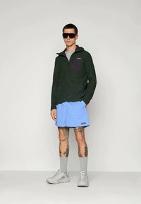 Zaļš kapuce ar rāvējslēdzēju un melnu krūšu kabatu, ko papildina gaiši zili šorti. Modelis valkā saulesbrilles un pelēkus apavus, demonstrējot kāju tetovējumus.