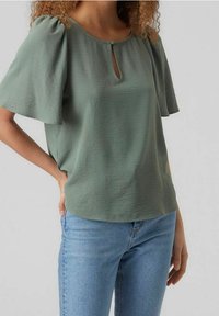 Blusa verde menta con maniche corte a onda, caratterizzata da un colletto a chiave. Tessuto morbido e vestibilità rilassata, abbinata a jeans azzurri chiari.