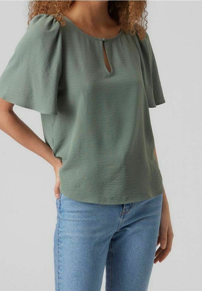Blusa verde menta con maniche corte a onda, caratterizzata da un colletto a chiave. Tessuto morbido e vestibilità rilassata, abbinata a jeans azzurri chiari.