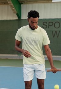 Hele roheline Nike t-särk graafilise disainiga, koos valgete tekstuursete lühikestega. Isik hoiab tennisereketit väljakul.