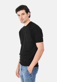 Maglietta nera a maniche corte con collo rotondo e taschino sul petto, realizzata in tessuto liscio, abbinata a jeans blu chiaro.
