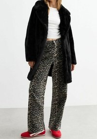 Černý kabát z umělé kožešiny přes bílé přiléhavé tričko, doplněný širokými kalhotami se vzorem leopardí kůže. Červené nazouvací boty doplňují outfit.