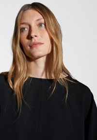 Top nero con scollatura ampia, caratterizzato da linee pulite e una texture liscia. Capelli lunghi e mossi parzialmente visibili, su uno sfondo neutro.