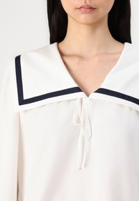 Hvit bluse med en stor marineblå krage i rektangulær form, V-hals og midtstående knytting. Myk tekstur og lett materiale.