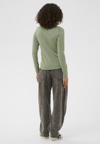 Ženska s kodrastimi lasmi oblečena v svetlo zeleno majico z dolgimi rokavi, ohlapne sivo-dolge jeans hlače in bele superge s črnimi črtami, obrnjena stran od kamere.