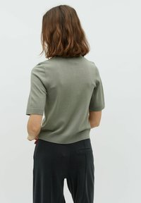 Korte mouwen trui in olijfgroen, met een aansluitend ontwerp, ribbelstiksel bij de halslijn en onderkant, gedragen met zwarte broeken. Zachte brei textuur.