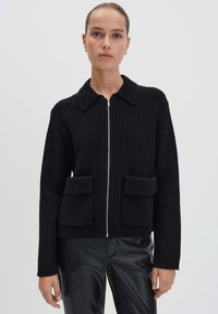 Vrouw in een zwart geribbeld vest met rits en kraag, voorzien van twee voorzakken met klep, gecombineerd met zwarte leren broeken, tegen een effen achtergrond.
