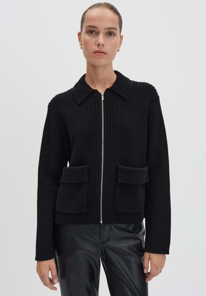 Woman wearing a black ribbed zip-up cardigan with collar and two front flap pockets, paired with black leather pants, against a plain background.