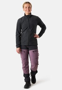 Giacca in pile nera con colletto alto, abbinata a pantaloni viola e neri con zip laterali e sezioni rinforzate sulle ginocchia.
