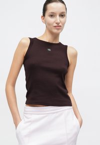 Σκούρο καφέ ριμπ φανελάκι με στρογγυλό ντεκολτέ και μικρό μαύρο λογότυπο στο κέντρο. Φορεμένο με ροζ ανοιχτόχρωμο ψηλόμεσο παντελόνι.