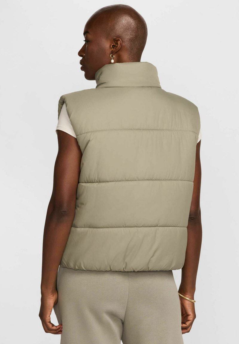 Persona con testa rasata che indossa un gilet trapuntato beige sopra una camicia bianca a maniche corte e pantaloni beige abbinati, in piedi di spalle su uno sfondo neutro.
