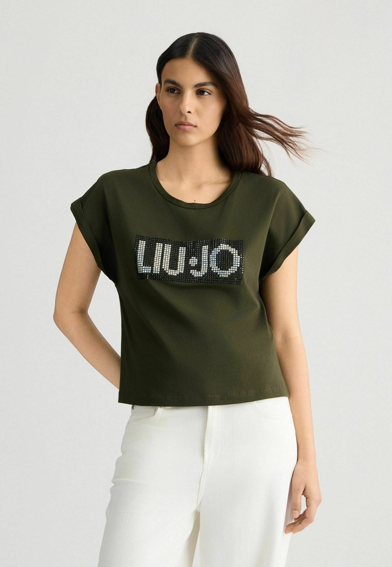 T-shirt cropped verde oliva con maniche arrotolate, caratterizzata da un logo "LIU·JO" impreziosito da paillettes su uno sfondo rettangolare nero.