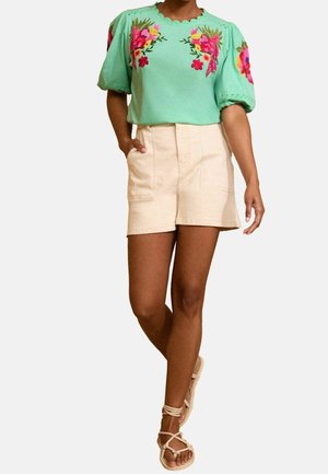 Vrouw draagt een muntgroene blouse met roze vogel- en bloemenborduurwerk, beige korte broek met hoge taille en beige veter-sandalen, staat met een been gekruist.