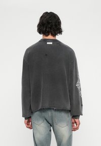 Persona con cabello rizado oscuro lleva una camisa de manga larga negra deslavada con detalles desgastados y jeans azul claro deslavado, de espaldas.