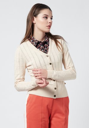 Cardigan in cotone beige con scollatura a V, caratterizzato da un motivo testurizzato e cinque bottoni scuri sulla parte anteriore, indossato sopra una camicia con motivo.