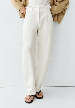 Person iført højttaljede vidde ben hvide bukser, beige jakke og sorte flipflops, stående mod en almindelig hvid væg.