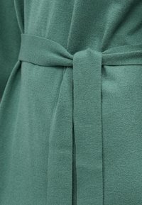 Grønt strikket stoff med en myk tekstur, med et bredt belte knyttet i midjen og subtile sømdetaljer på sidene.