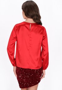 Blusa rossa in satin a maniche lunghe con apertura a forma di fessura sul retro, abbinata a una mini gonna bordeaux tempestata di paillettes.