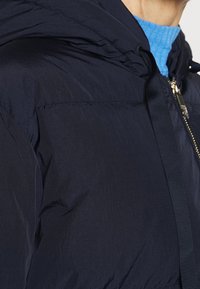 Námořnická prošívaná bunda s kapucí, vyrobená z texturované látky, s předním zipem a tónovanou stuhou podél zipu a límce.