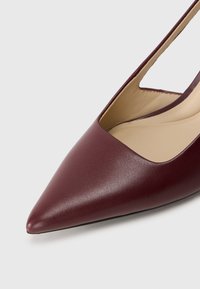 Scarpa piatta in pelle bordeaux con punta appuntita e cinturino slingback, con fodera beige su una superficie bianca.