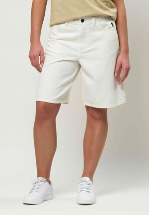 Person iført hvide denim knæshorts, beige top og hvide sneakers, stående mod en ensfarvet baggrund.