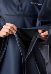 Marinblå vattentät poncho med justerbar hals, framficka och ett mjukt innerfoder. Har en slät yta och kontrasterande svarta detaljer.