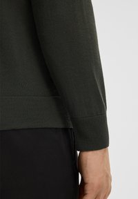 Dunkelgrüner Strickpullover mit geripptem Bündchen, glatter Textur und leicht zulaufendem Saum, kombiniert mit schwarzem Stoff darunter.