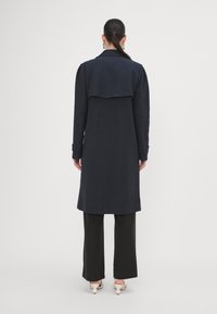 Largo abrigo azul marino con una parte posterior tipo capa, con un cuello entallado, mangas largas con detalles en los puños, combinado con pantalones negros acampanados.
