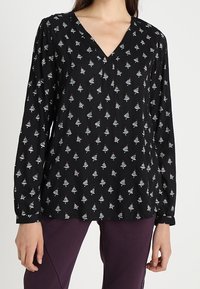 Blouse noire à manches longues avec un col en V, présentant un petit motif d'arbres en blanc et bleu clair, fabriquée en tissu léger.