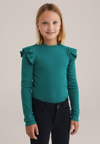 Fiatal lány, aki szőke hajú, teal színű, hosszú ujjú pólót visel ráncos vállakkal és fekete nadrággal, egy sima háttér előtt áll.