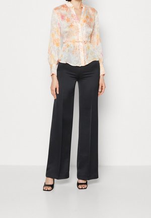 Femme portant un chemisier floral transparent avec un lien à la taille, un pantalon noir à jambe large et des sandales à talons ouverts noires, se tenant devant un fond blanc.