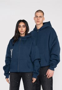 Sweatshirts à capuche zippés bleu marine en tissu doux, avec une coupe oversize, de grandes poches et un logo brodé sur le côté gauche.