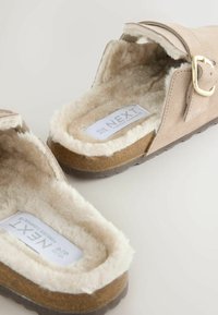Zapatillas de ante beige con forro de piel sintética, con apertura trasera y detalle de hebilla en tono dorado. Suela gruesa de goma marrón, diseño plano para mayor comodidad.