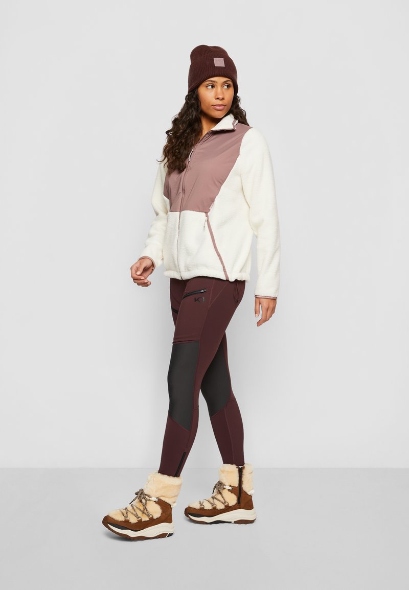 Leichte Fleecejacke in Creme und Mauve, kombiniert mit burgunderfarbenen Leggings mit schwarzen Akzenten. Braune Stiefel mit Futter aus Fell vervollständigen das Outfit.