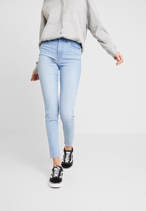 Person iført lyseblå skinny jeans, grå hættetrøje og sort-hvide sneakers, der går fremad mod en ensfarvet baggrund.