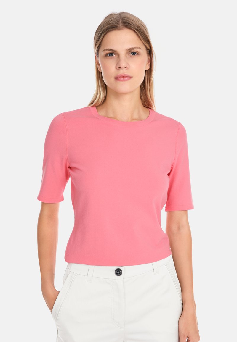 Short-sleeve, round-neck top in bright pink, made of smooth fabric. Paired with white pants featuring a button detail.