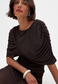 Black draped blouse with gathered details, featuring soft jersey fabric. Model wears a white bracelet and large gray hoop earrings.