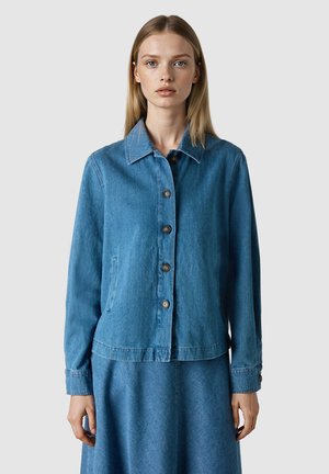 Mujer joven con chaqueta de mezclilla azul abotonada y falda a juego, posando contra un fondo gris claro liso.