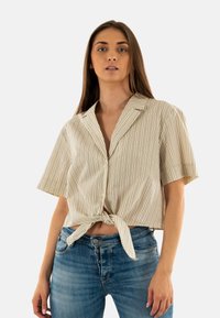 Camicia a righe, a maniche corte, con colletto in beige e crema, con un dettaglio annodato davanti. Abbinata a jeans in denim blu a vita alta.